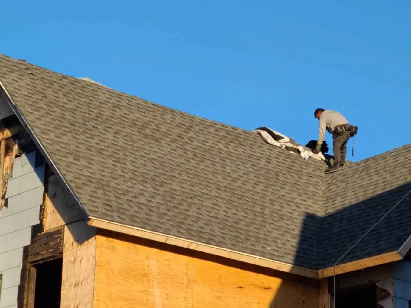 A worker on a roof installing shingles during daytime with a clear blue sky in the background.
