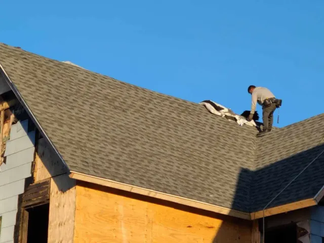 A worker on a roof installing shingles during daytime with a clear blue sky in the background.