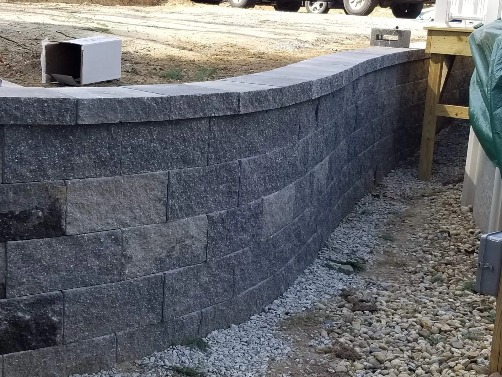 A curved stone retaining wall made of interlocking gray granite blocks, with a gravel pathway beside it. There is construction material and a wooden post with a tarp on the right side.