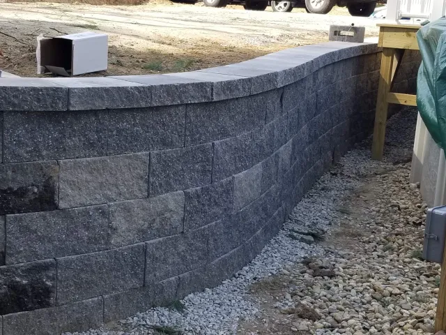 A curved stone retaining wall made of interlocking gray granite blocks, with a gravel pathway beside it. There is construction material and a wooden post with a tarp on the right side.