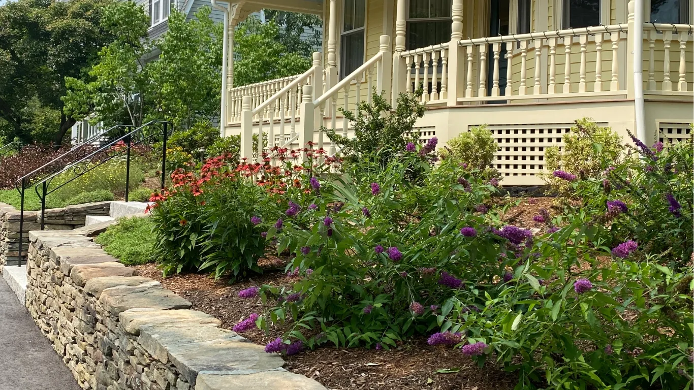 A garden with purple and red flowers borders a stone retaining wall. Behind it is a house with a light yellow wooden porch featuring ornate railing details. A paved pathway with metal handrails leads up to the porch.