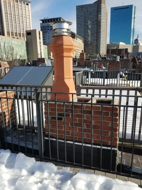 Red brick chimney on a rooftop with snow, black railing, city buildings in the background.