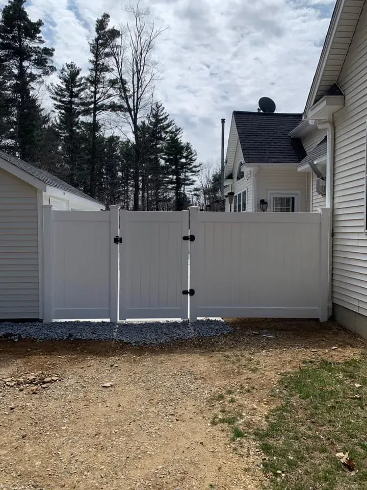 A white vinyl privacy gate stands between a garage and a house, situated on a dirt driveway with trees in the background.