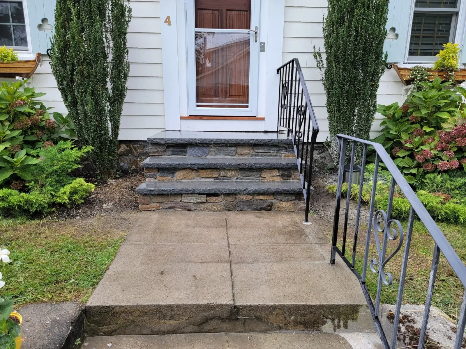 Stone front steps lead to a white door, framed by tall narrow evergreens, hydrangeas, and window boxes