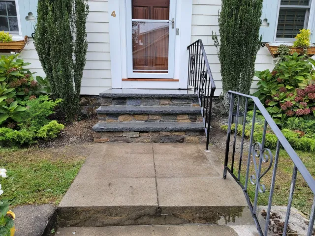 Stone front steps lead to a white door, framed by tall narrow evergreens, hydrangeas, and window boxes