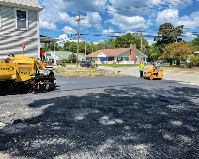 Asphalt paving in progress on a street. A yellow paver and roller flatten the fresh blacktop.