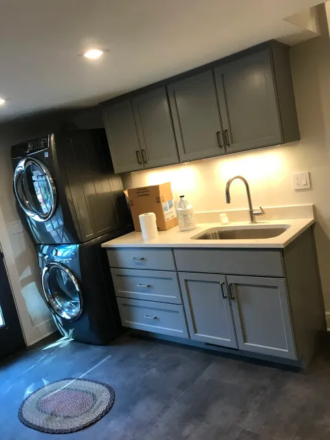Laundry room with stacked black washer and dryer units, gray cabinets, a white countertop with cleaning supplies, a paper towel roll, a sink, and a rug on the dark floor.