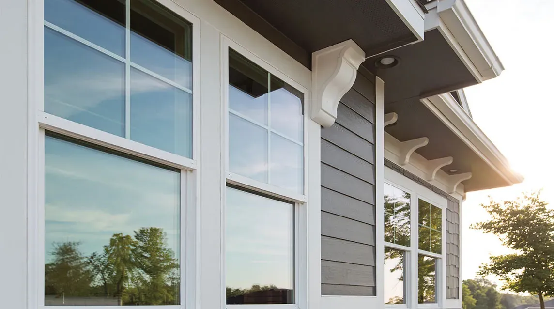 Close-up of white framed windows on a gray house exterior reflecting trees and sky at sunset.