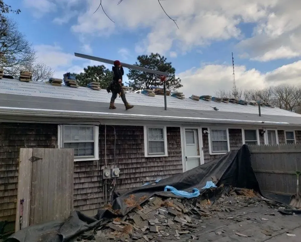 Roofer carries a metal beam across a roof; a pile of shingles and debris in the foreground.
