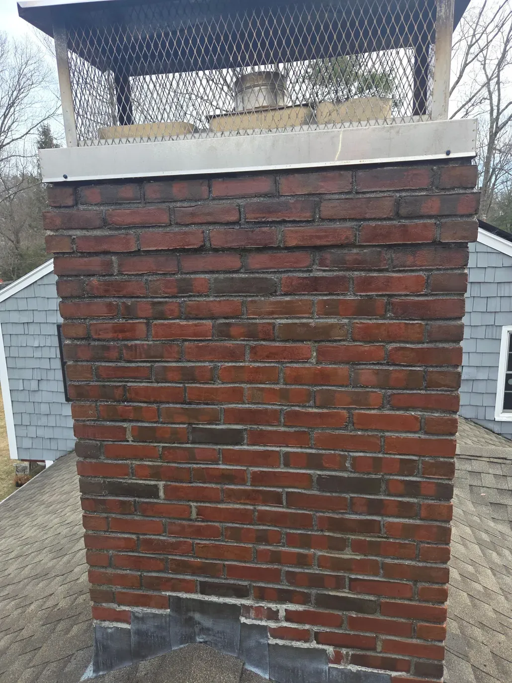 Brick chimney with a metal cap and white trim on a shingled roof, viewed from outside.