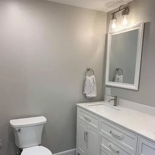 A modern bathroom with light gray walls, a white toilet, a white vanity with drawers, a rectangular mirror above the sink, and a wall-mounted towel ring holding a patterned hand towel. Two lights are above the mirror.