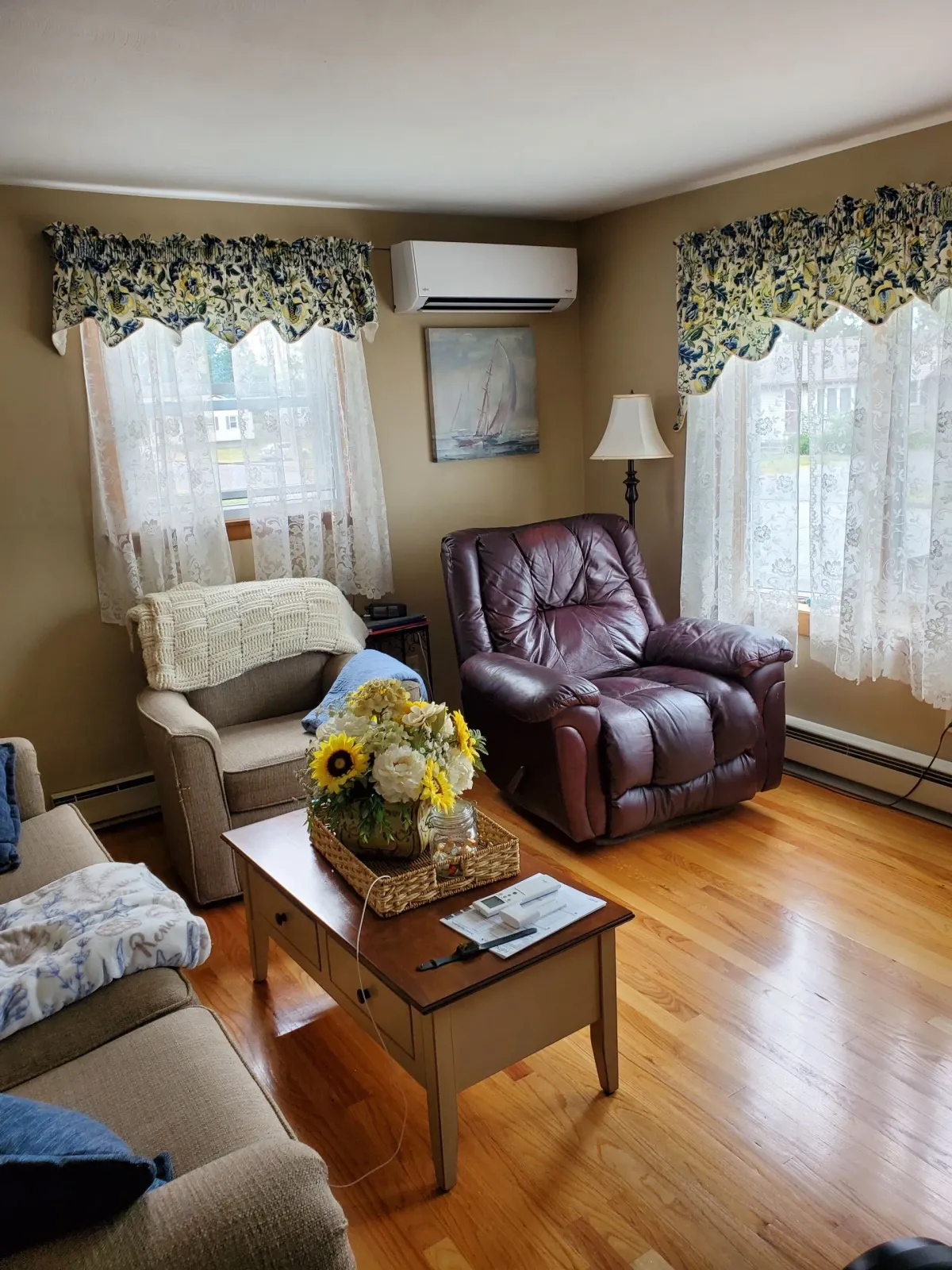 Living room with hardwood floors, two windows with lace curtains, a leather recliner, and a beige sofa.