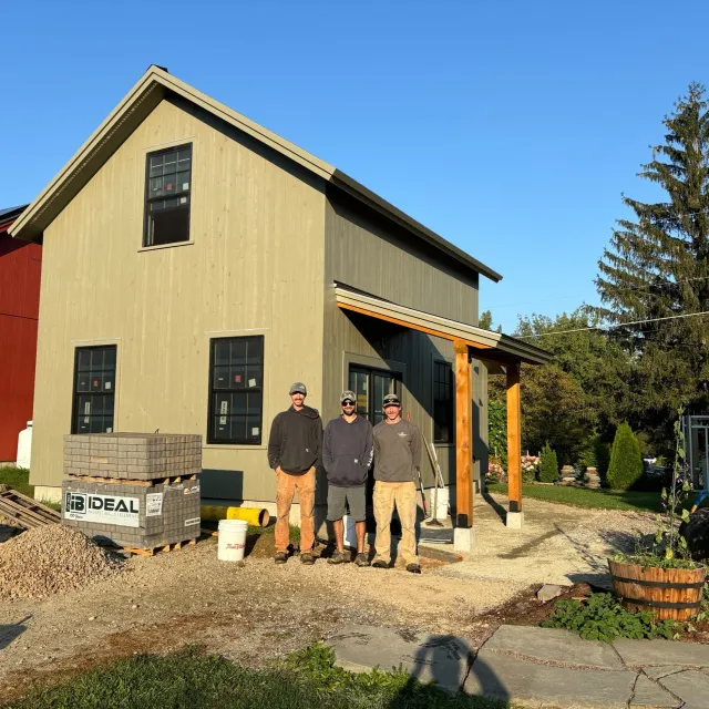 Three men standing together in front of a house under construction, with construction materials and tools around them, during daylight.