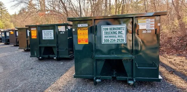 Several dark green dumpsters lined up on a gravel lot near trees.