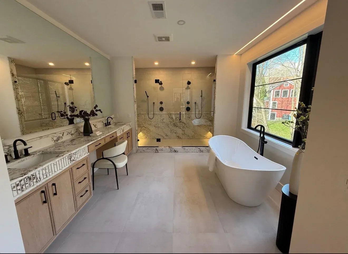 Modern bathroom with a large window, freestanding bathtub, marble vanity with two sinks, and a walk-in shower with glass walls, dark fixtures, and built-in lighting.