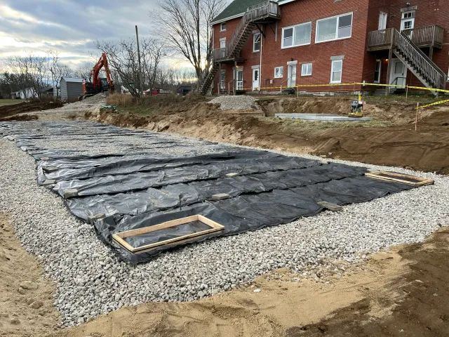 Excavated foundation pit with geotextile membrane, gravel base, and wooden frames near brick building