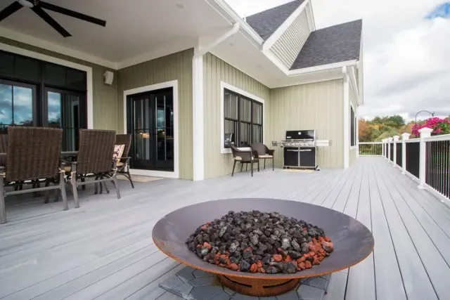 Outdoor patio with fire pit, gray deck, green house exterior, black windows, and grill.