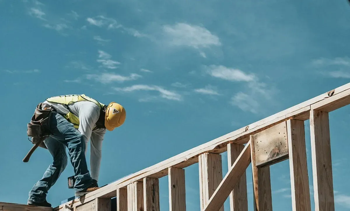 Construction worker wearing a yellow hard hat and vest working on a wooden house frame under blue sky.