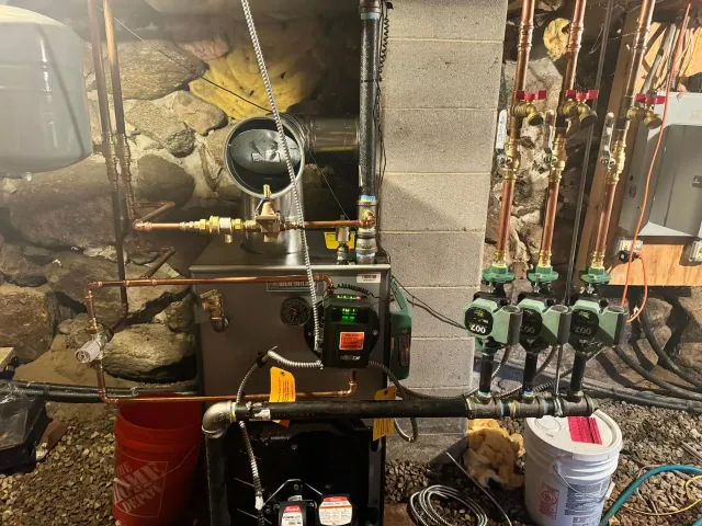 Basement utility area with pipes, valves, gauges, and storage bins against a stone wall.