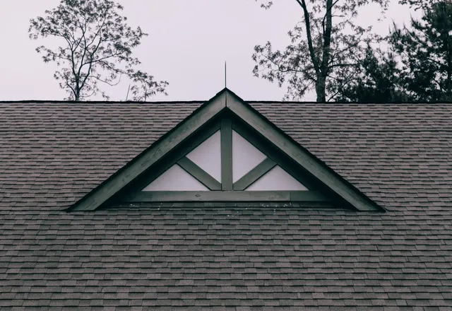 Peaked roof gable with triangular window and spire against gray sky and trees