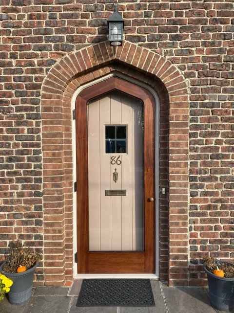 Front door with house number 86, set in a brick wall with an arched brick frame. Two black planters with pumpkins and flowers on either side of the door, a black doormat in front, and a black outdoor lantern above.