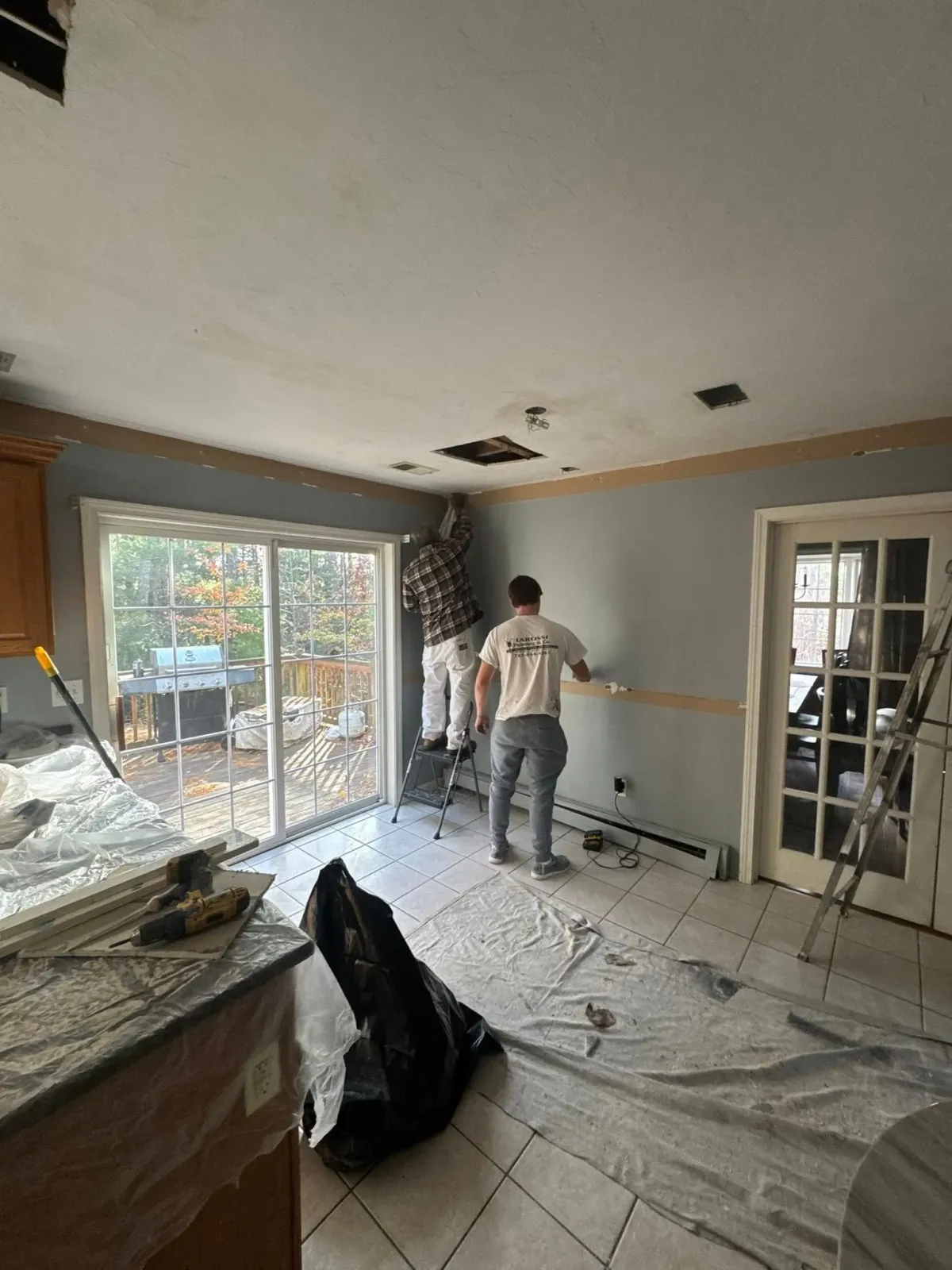 Worker installing wall trim in a messy living room under renovation, with plastic sheeting and exposed ceiling patches.