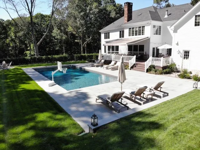 Backyard with white house, swimming pool, lounge chairs, umbrellas, and surrounding greenery.