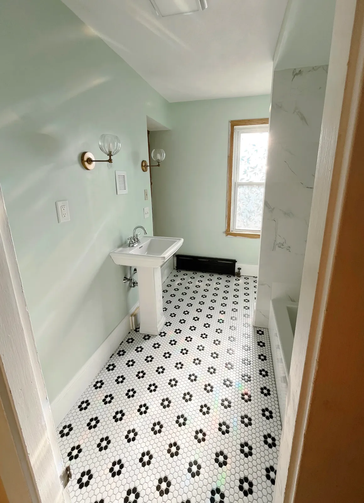 A small bathroom with a green wall, a white pedestal sink, black and white floral patterned tile floor, two wall-mounted light fixtures, a window with wooden trim, and a partial view of a bathtub with a marble-looking wall surround.