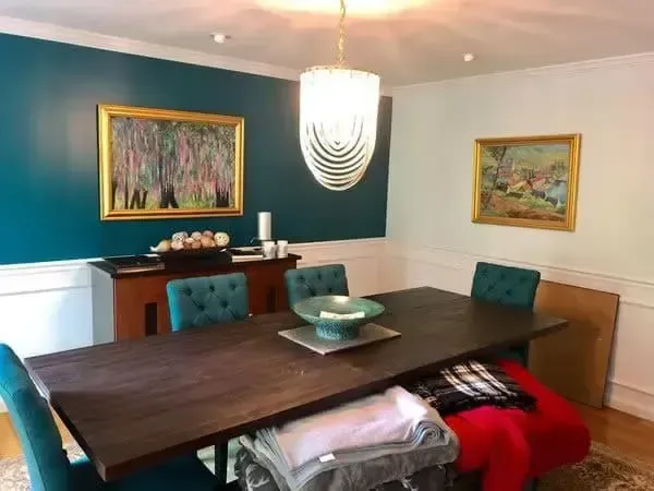 Dining room with teal walls, dark wood table, tufted chairs, hanging light, and framed paintings.
