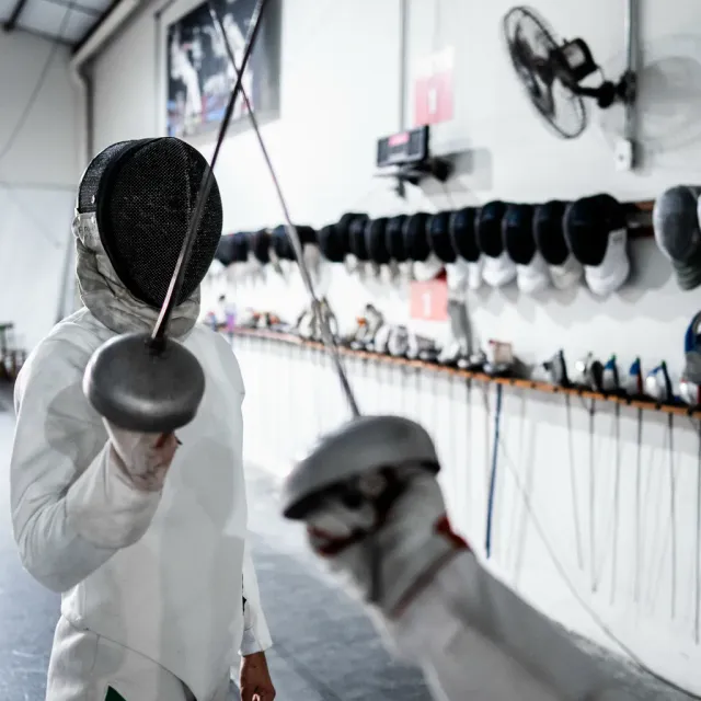 Two épée fencers sparring during class practice at Allez Fencing Club in Watertown, MA, with fencing gear and equipment visible in the background.