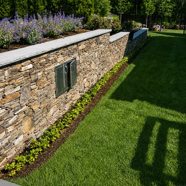 Stone retaining wall with drainage access, flowering plantings and fresh green lawn
