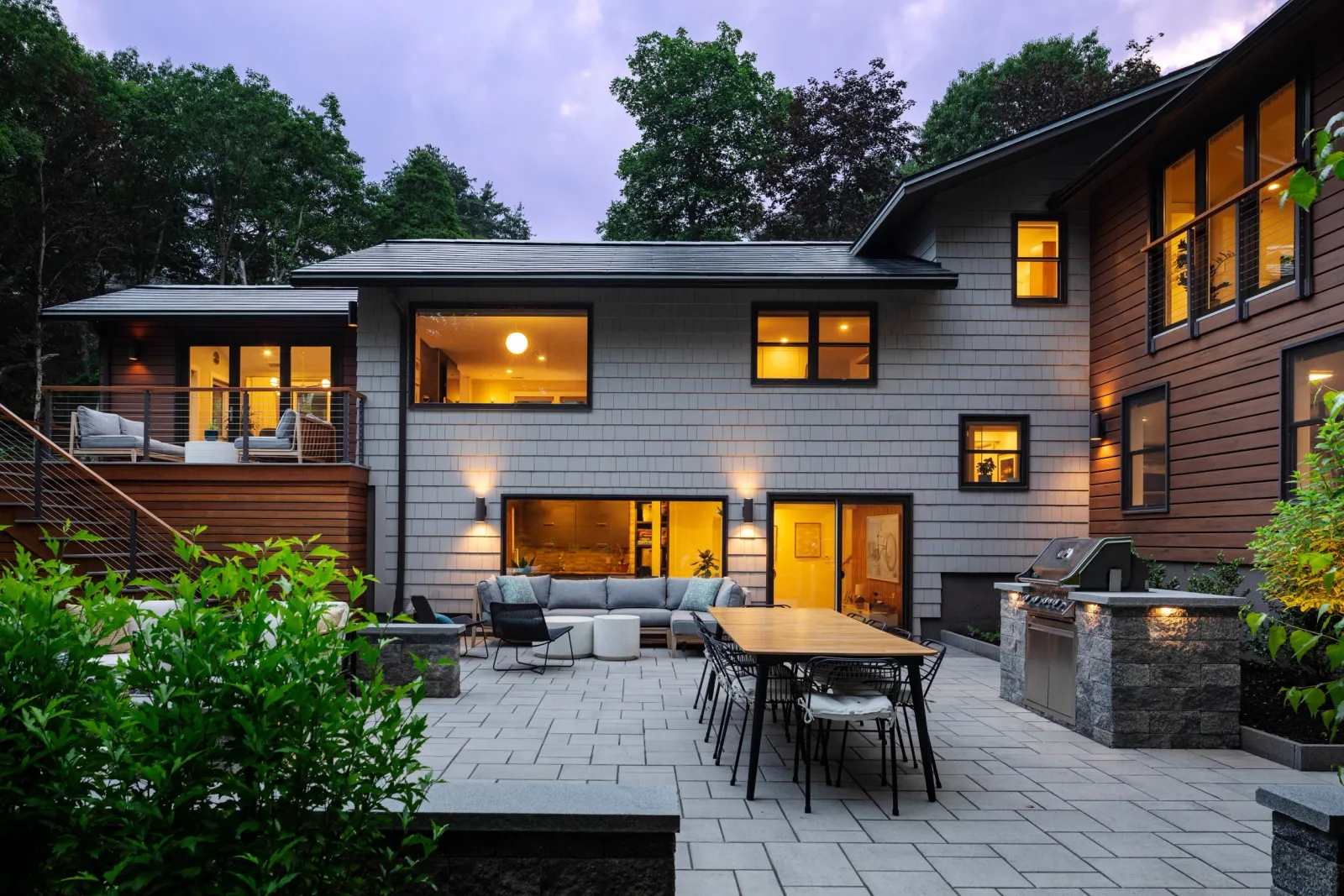 Modern two-story house with a large outdoor patio, lit interior windows, outdoor seating, dining table, and a built-in grill, surrounded by trees at dusk.