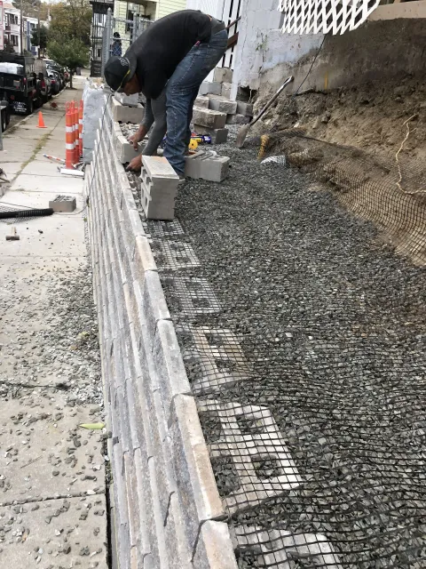 Man building a retaining wall on a city sidewalk with gray bricks and gravel.