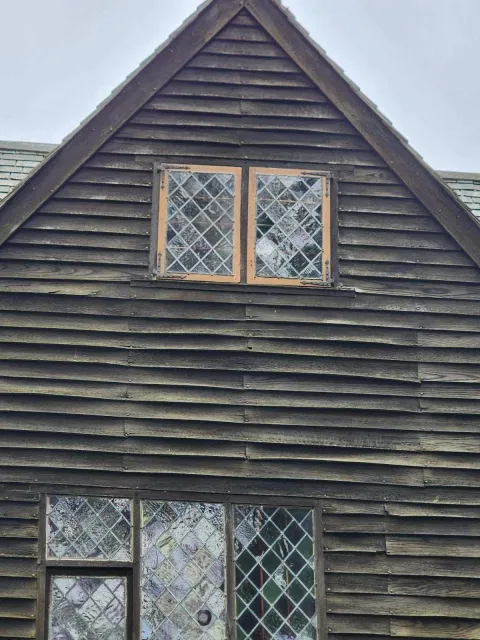 A wooden house with boarded-up diamond-pane windows on the upper level and a ground-floor window with many small diamond panes, all with weathered wood siding.