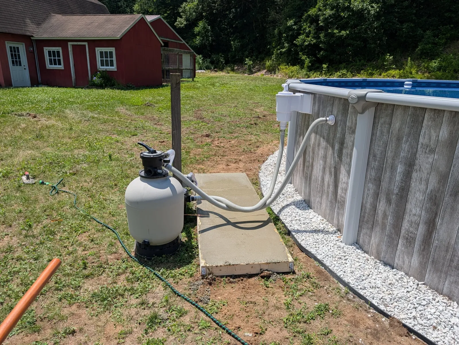 An above ground swimming pool with a white exterior and blue interior water, connected to a filter system, on a concrete pad in a backyard. There is a red barn with white trim and several windows in the background, surrounded by green grass and trees