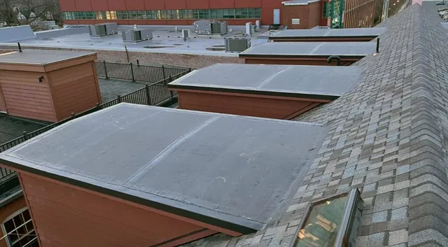 Chris Battaini Roofing - Rooftop view of several small structures with flat gray roofs adjacent to a larger building. The shingles are gray, and the larger building has a red exterior.