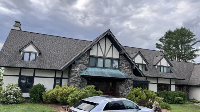 Tudor style home with stone and stucco facade, dark grey shingle roof, dormers and a brick chimney