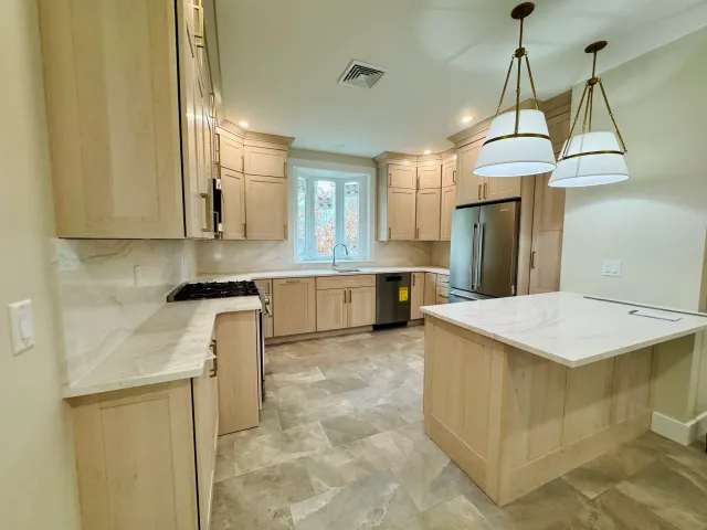 Kitchen with light wood cabinets, marble countertops, stainless steel refrigerator, gas stove, and pendant lighting over a central island.