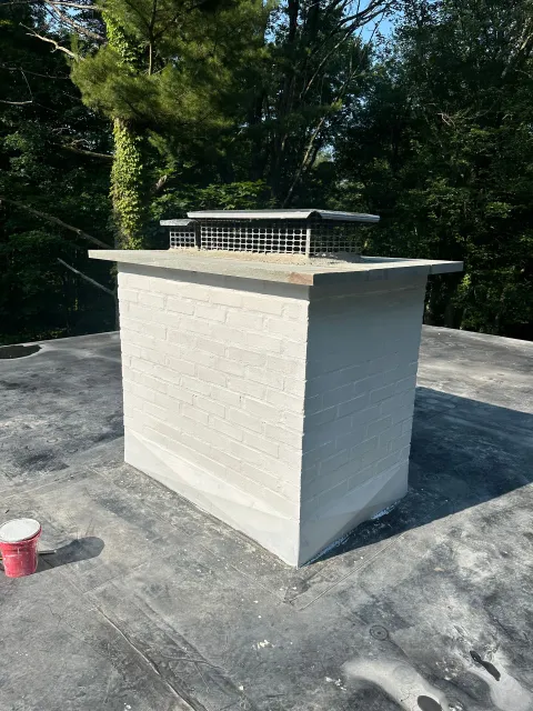 A large, rectangular concrete chimney with a metal cap stands on a flat, dark gray roof surrounded by trees.