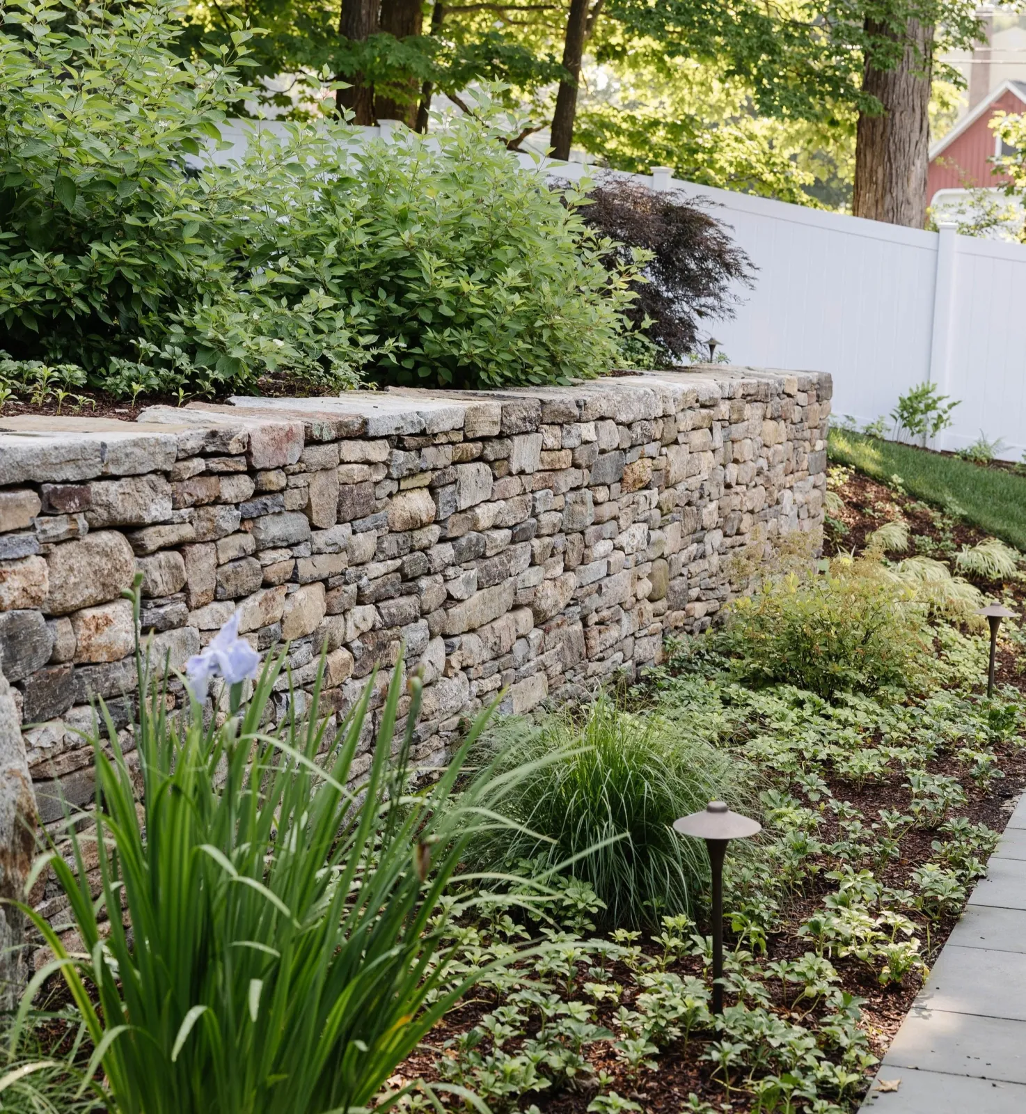 A landscaped backyard garden with a stone retaining wall, green shrubs, flowering plants, and a white fence in the background.