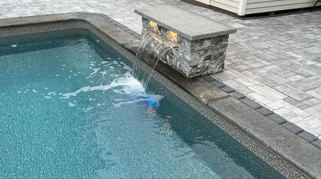 A swimming pool with water flowing from a stone waterfall feature, surrounded by stone and brick paving.