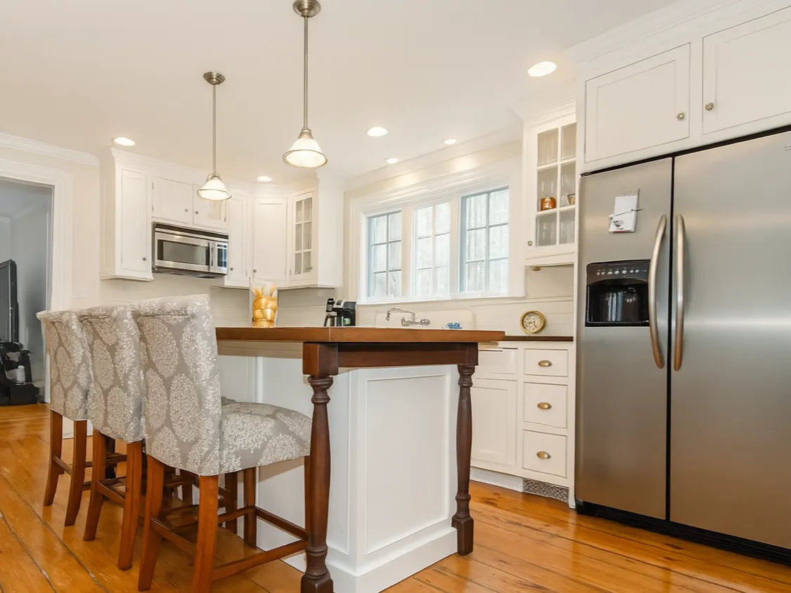 Modern kitchen with white cabinets, black countertops, stainless steel appliances, and a white subway tile backsplash. There is a red kettle on the stove, a vase with pink flowers on the counter, and decorative blue vases near the window. Natural light streams in through the window.