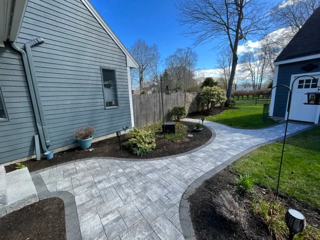 A backyard patio with a curved gray stone walkway, green grass, and a blue house with siding. There are several bushes, small trees, garden lights, and a wooden fence in the background. The sky is partly cloudy with some sunshine.