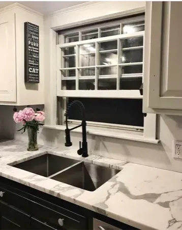 Modern kitchen sink with black faucet, white cabinets, and window over marble countertop