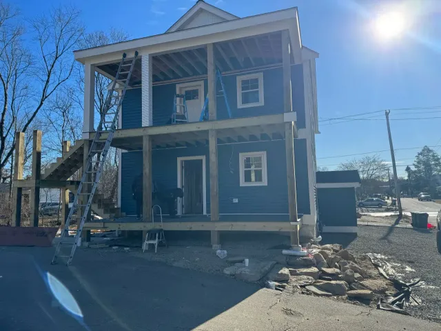 Two-story blue house under construction with wood decks and a ladder, on a sunny day.
