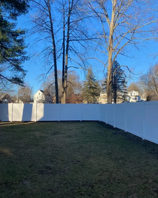 View of a backyard with a white privacy fence, leafless trees, and houses in the background under a clear blue sky during winter.