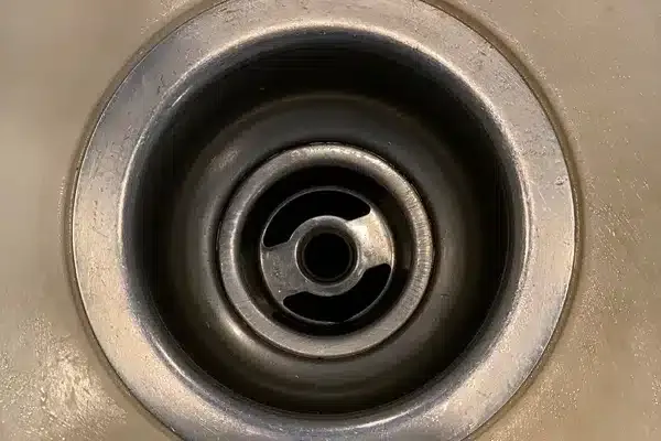 A close-up of a metal sink drain with a circular strainer in a beige countertop.