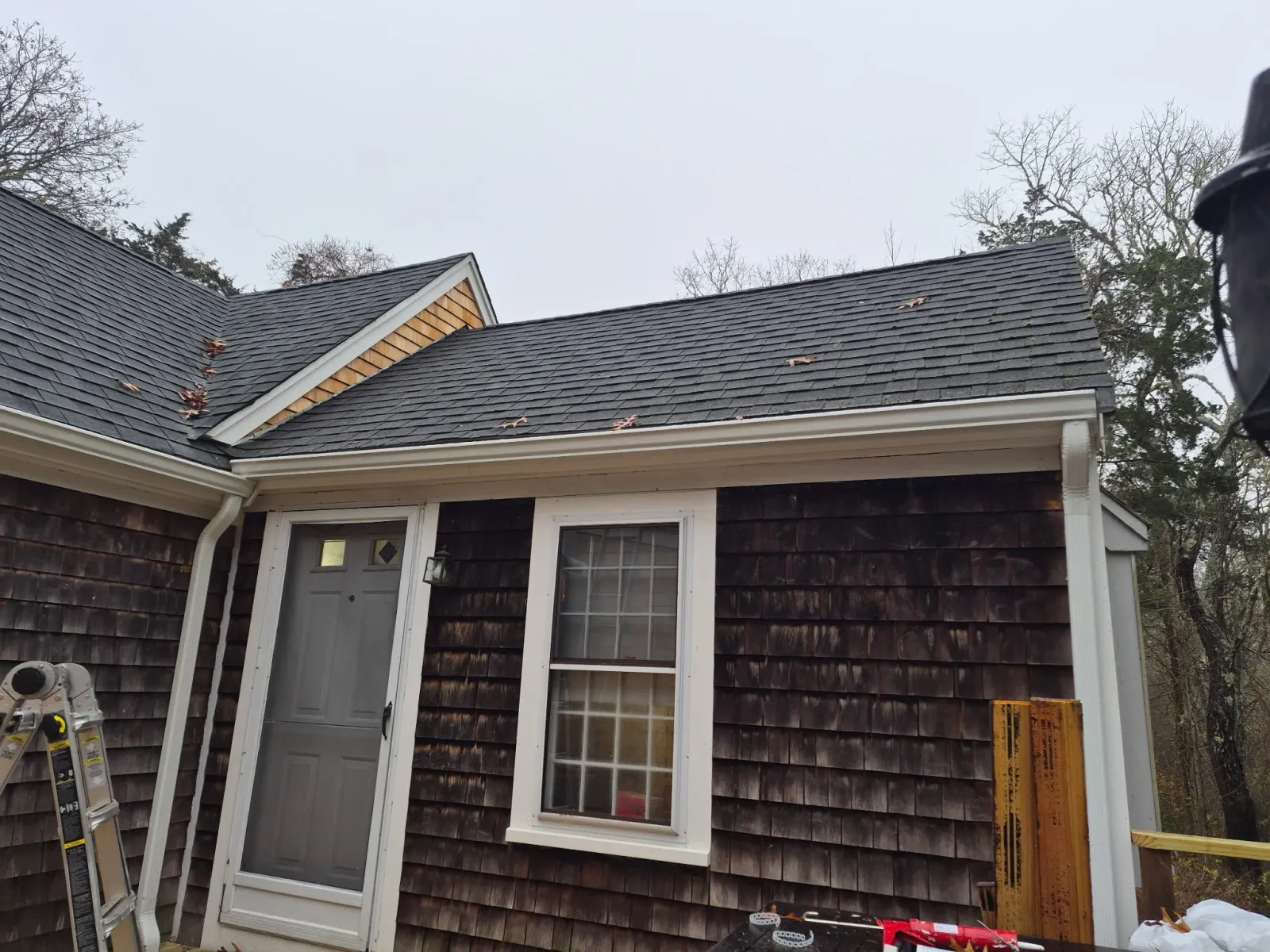 Exterior of a house with dark wooden shingles, a white door with a small window, and a window next to it. The roof has gray shingles with a few fallen leaves, and there is a ladder and construction tools nearby. The sky is overcast and trees are in t