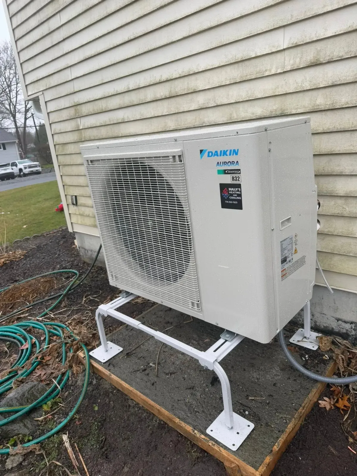 A Daikin outdoor HVAC unit sits on a white metal stand on a concrete pad next to the tan vinyl siding of a house.