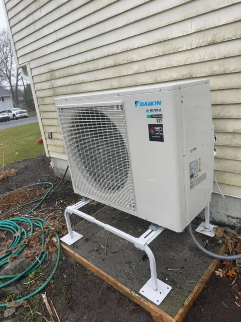 A Daikin outdoor HVAC unit sits on a white metal stand on a concrete pad next to the tan vinyl siding of a house.